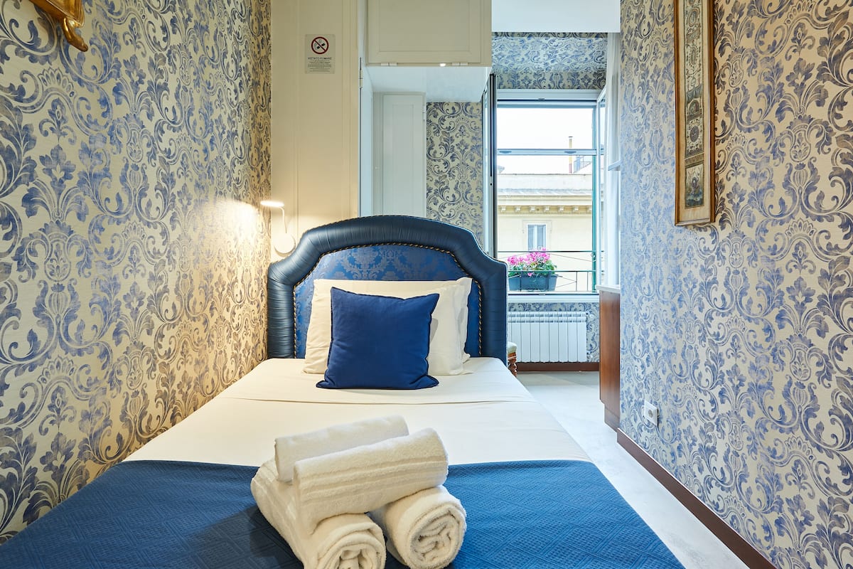 A hotel room with a bed, blue and white patterned wallpaper, a blue headboard, and a view of the outside balcony.