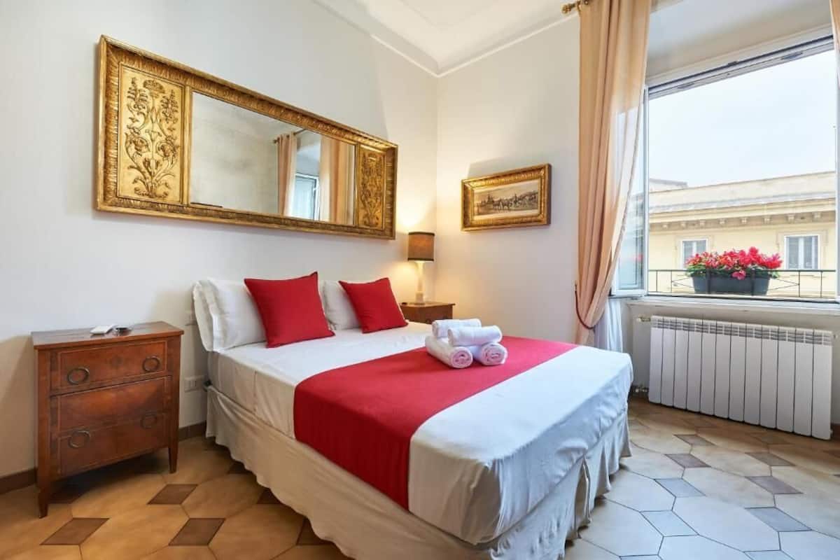 A hotel room with a large bed, a nightstand, a dresser, a window with a view of buildings and flowers, and a large mirror on the wall.