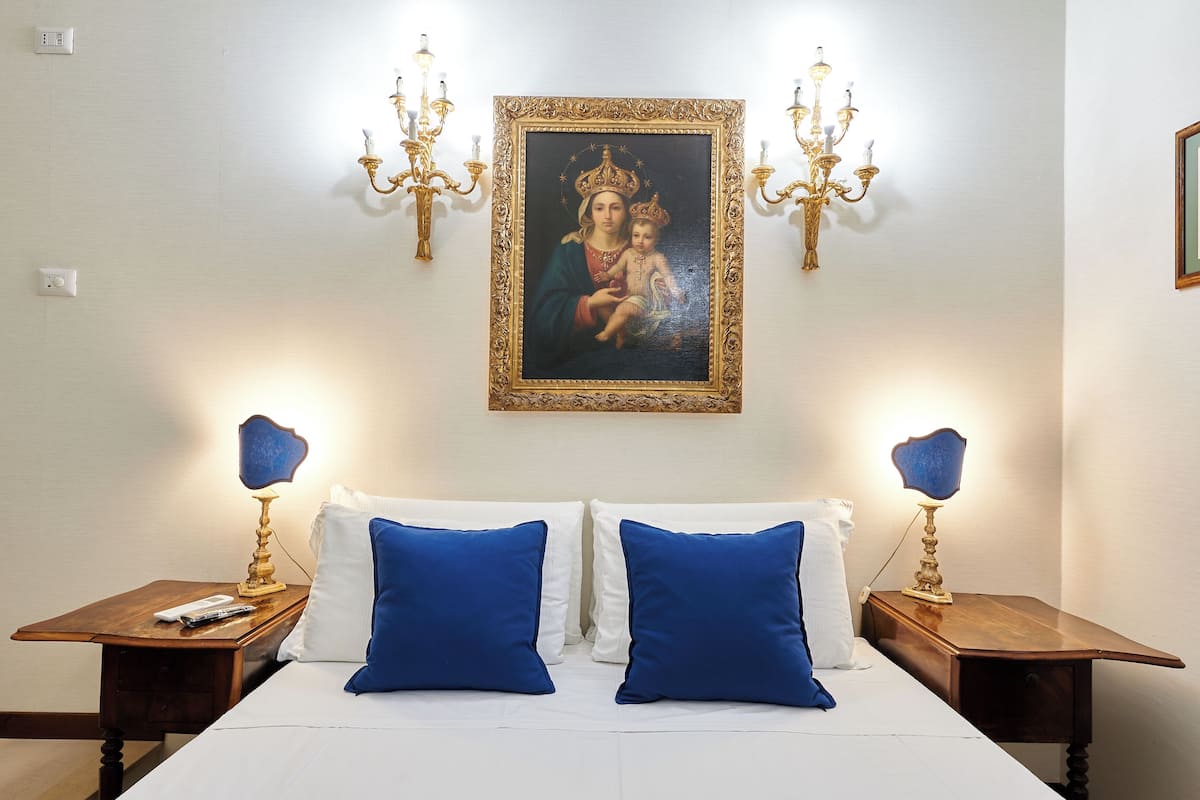 A hotel room with a bed, two nightstands, a chandelier, and a framed religious painting on the wall.