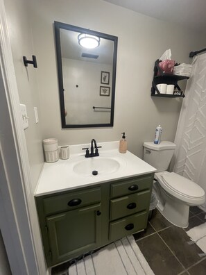 Un baño con un lavamanos blanco, un mueble de baño verde y un inodoro. Hay un espejo sobre el lavamanos y una repisa con artículos de tocador.