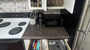 A compact kitchen with a white stove, black microwave, and stainless steel refrigerator. A drawer with utensils is visible, and a cabinet above contains various kitchen items. - Cozy 1BR Apt Minutes From Skyline Drive!! (Amissville)