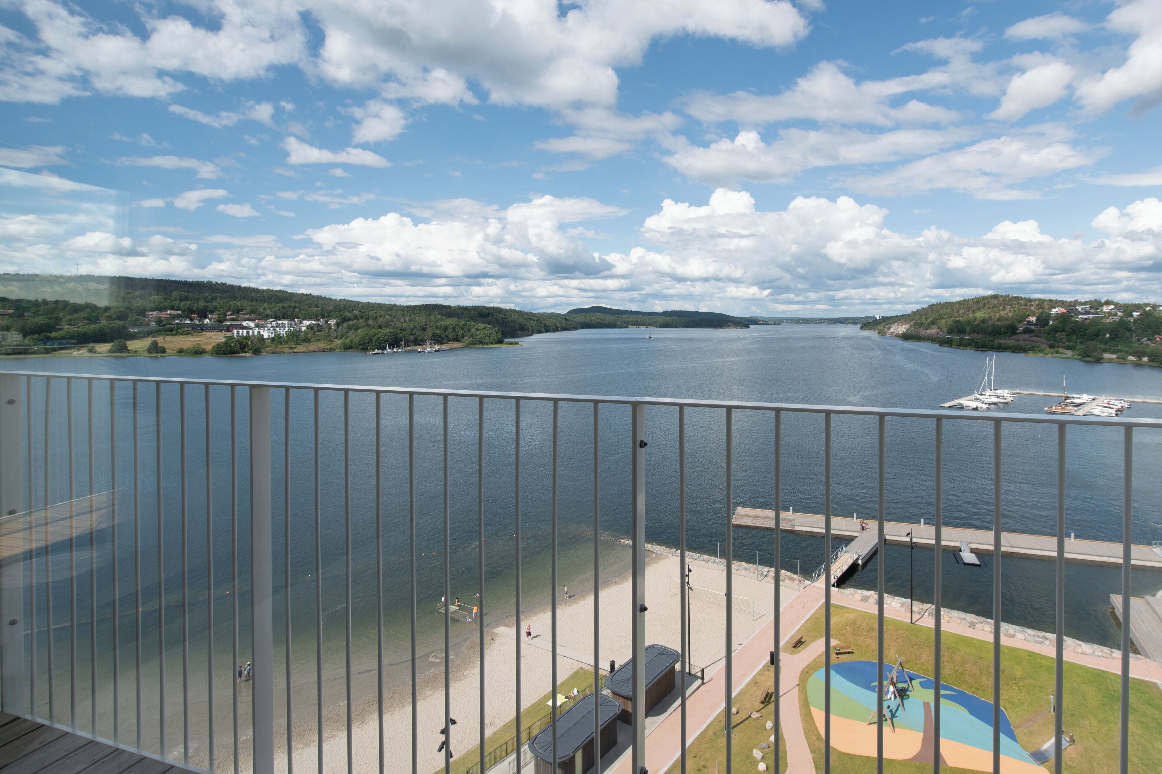 En utsikt från en balkong med utsikt över en sjö, en småbåtshamn, en strand och en lekplats.