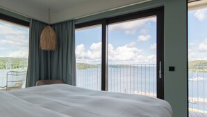 Habitación de hotel con una cama grande, una silla y vistas a un lago y montañas a través de amplias ventanas.