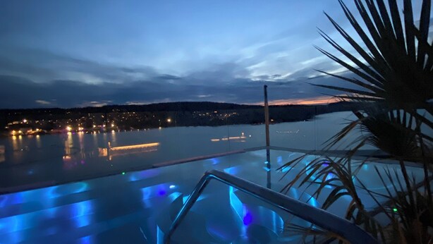 Una piscina infinita con iluminación azul al anochecer, rodeada de palmeras y un paisaje urbano al fondo.