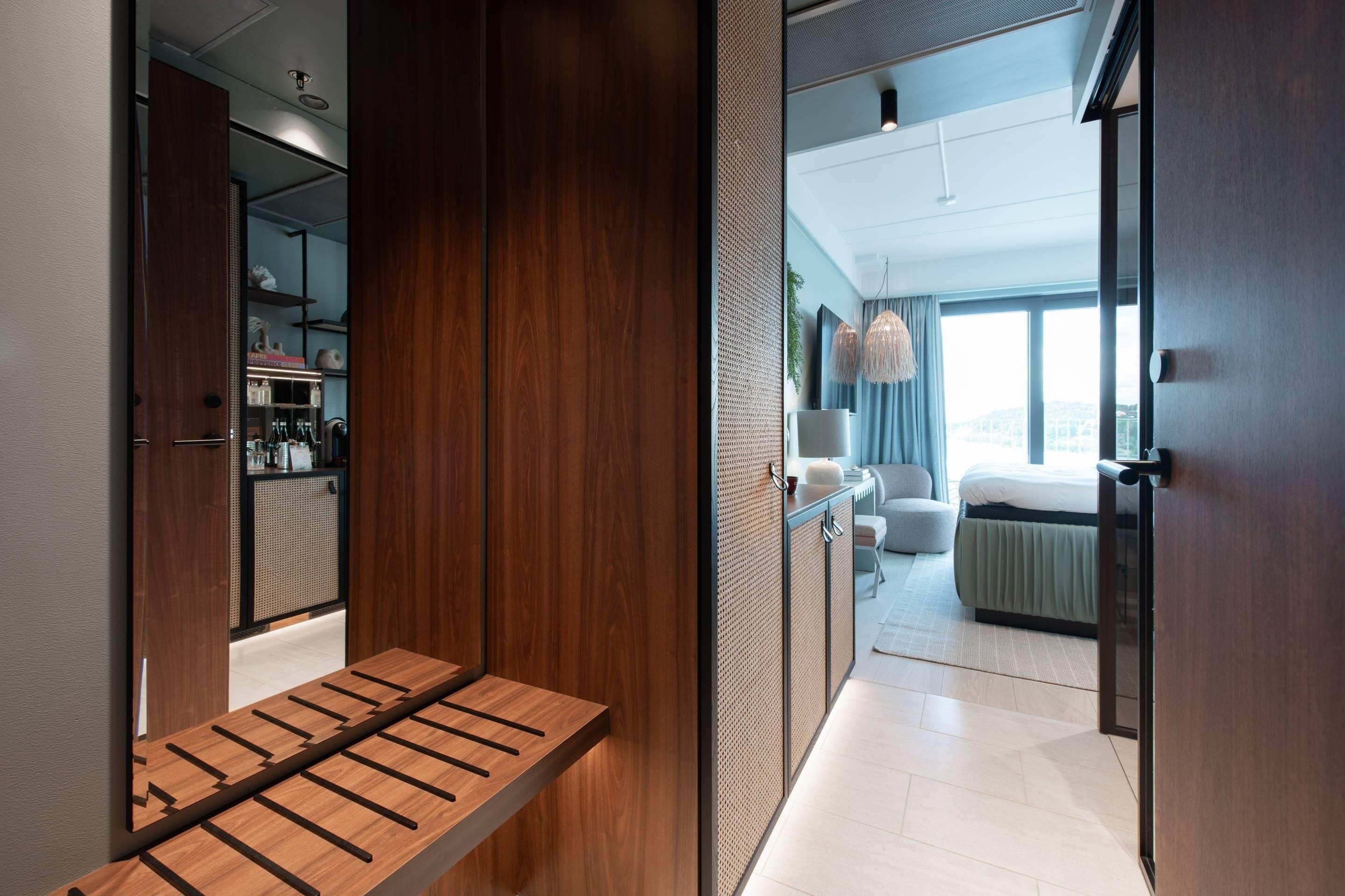 A modern hotel room with a wooden bench, a kitchenette, and a view of the city.