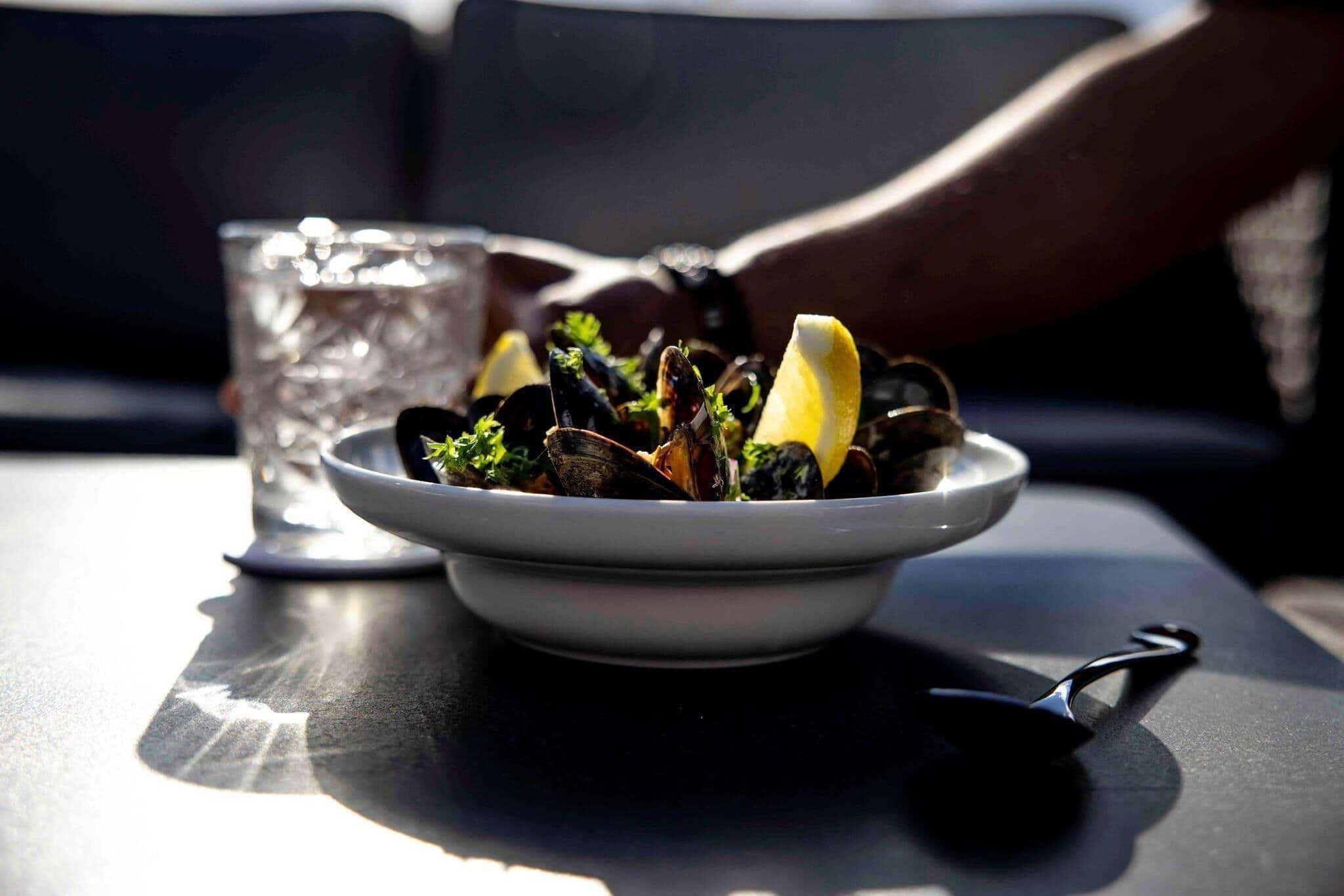 Un plato de mejillones con gajos de limón sobre una mesa, junto a un vaso de agua y una cuchara.