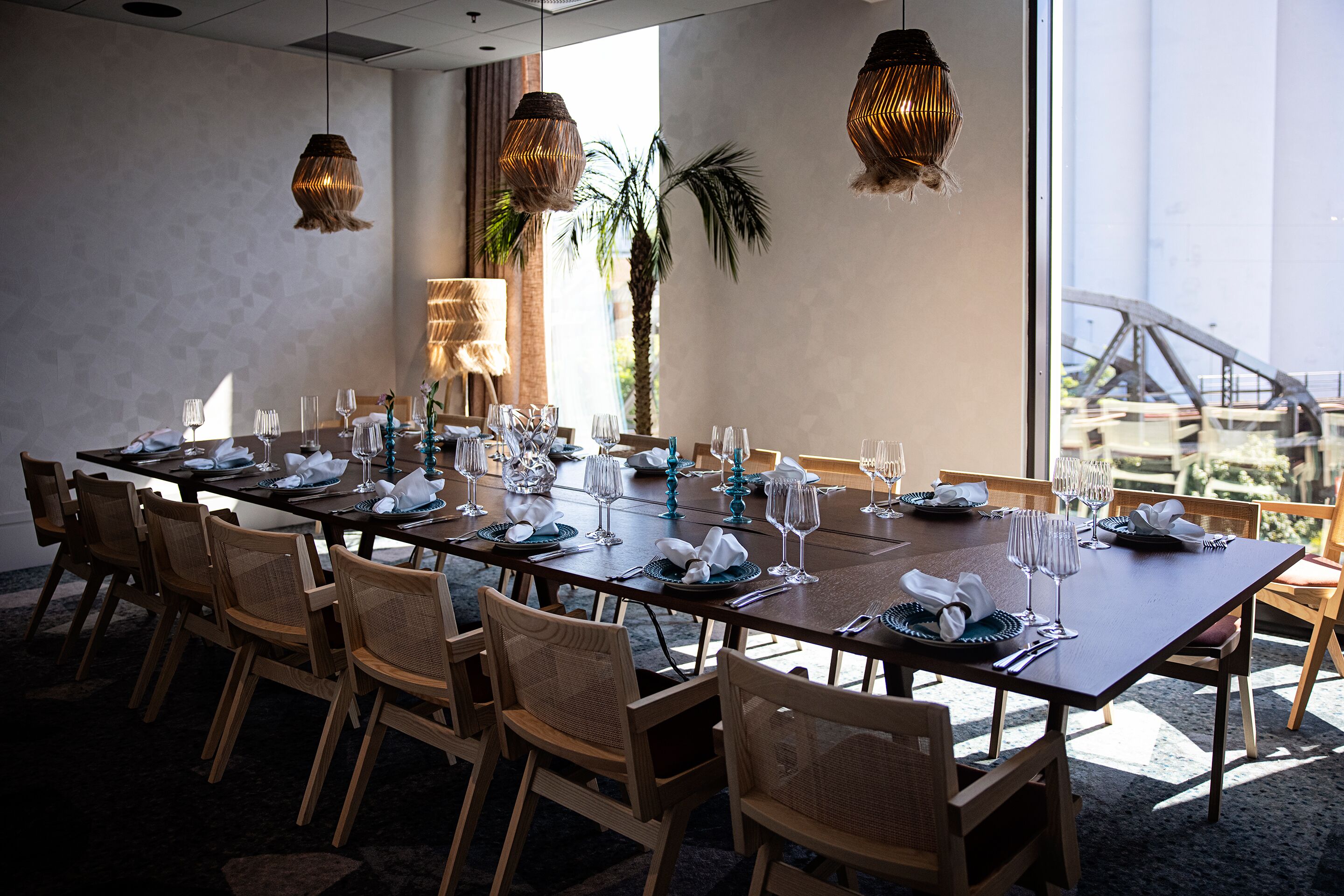 A dining area with a long table set for a meal, surrounded by wicker chairs, and large windows offering a view of industrial structures.