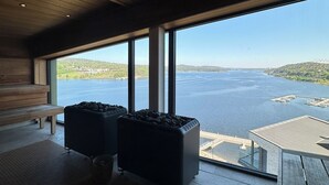 Un interior moderno con techo de madera, ventanas amplias que ofrecen vista a un lago y un paisaje distante, y una zona de estar con un banco y dos otomanas negras.