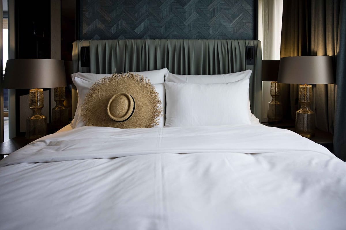 A neatly made bed with white linens, a straw hat on one pillow, and two lamps on either side.