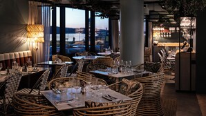 Un restaurante con mobiliario de mimbre, mesas dispuestas con copas de vino y cubiertos, y vistas a un cuerpo de agua a través de amplios ventanales.