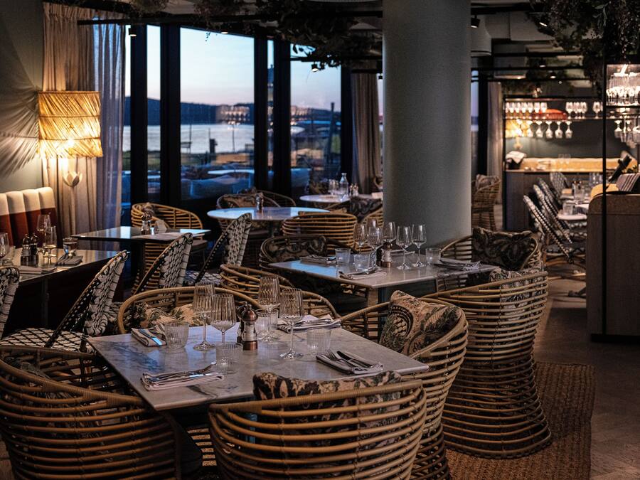 A restaurant with wicker furniture, tables set with wine glasses and cutlery, and a view of a body of water through large windows.