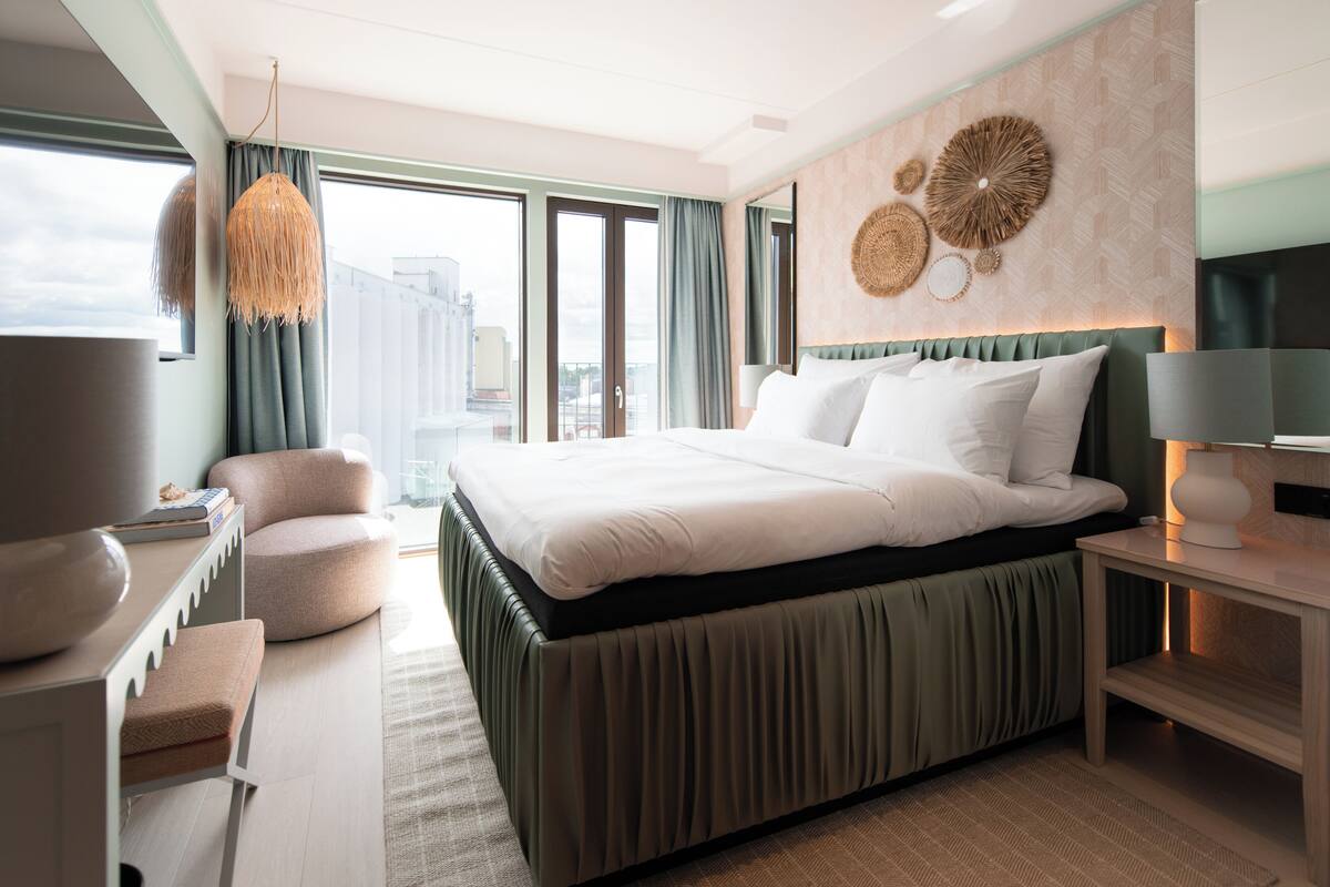 A modern hotel room with a large bed, a desk, a chair, and a view of the city.