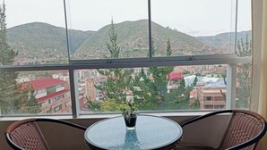 Ein Balkon mit Korbstühlen und einem Glastisch, mit Blick auf eine Stadtlandschaft mit Gebäuden und Hügeln.
