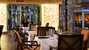 Um restaurante com parede de pedra e lareira. Mesas montadas com taças de vinho e talheres.