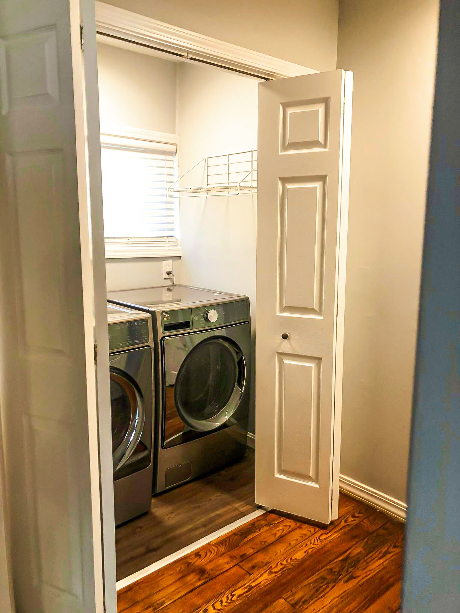 A laundry room with two stacked front-loading washers and dryers, a window with blinds, and a door leading to another room.