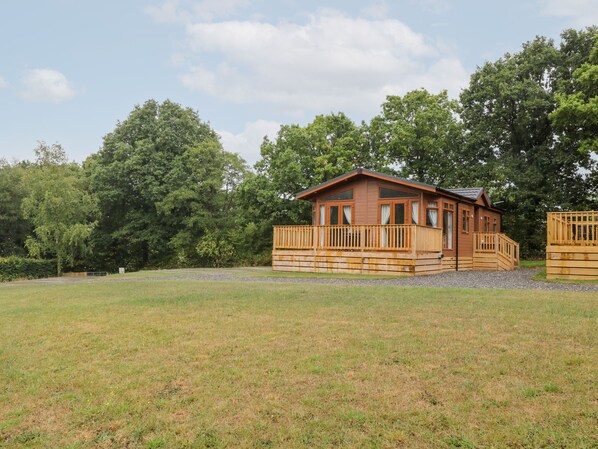 A wooden cabin with a balcony, surrounded by a grassy field and dense trees. - Woodpecker Lodge (Beaworthy)
