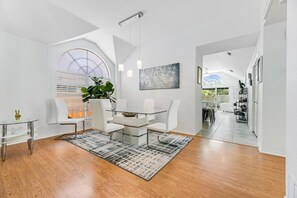 Una sala moderna con una mesa de comedor de vidrio, sillas blancas, un ventanal con persianas, una planta en maceta y un cuadro en la pared.