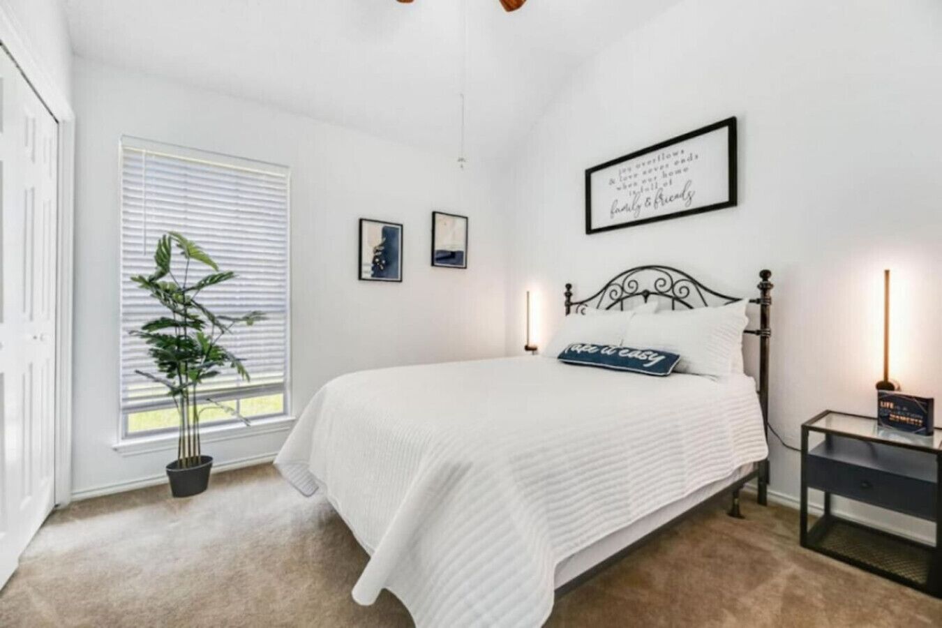 A bedroom with a bed, a bedside table, a nightstand, a potted plant, and a window with blinds.