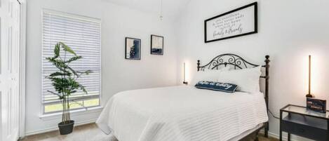 A bedroom with a bed, a bedside table, a nightstand, a potted plant, and a window with blinds.