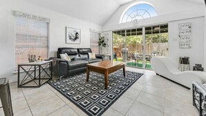 Amplia sala de estar con un sofá de cuero negro, una mesa de centro de madera y un tapete estampado. Hay un ventanal con persianas y vistas al exterior.
