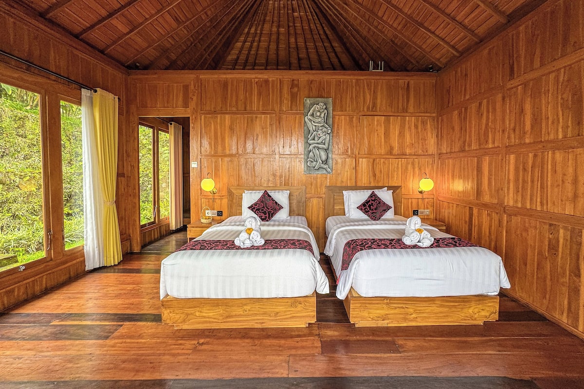 A wooden room with two beds, a wooden ceiling, and a view of greenery outside.