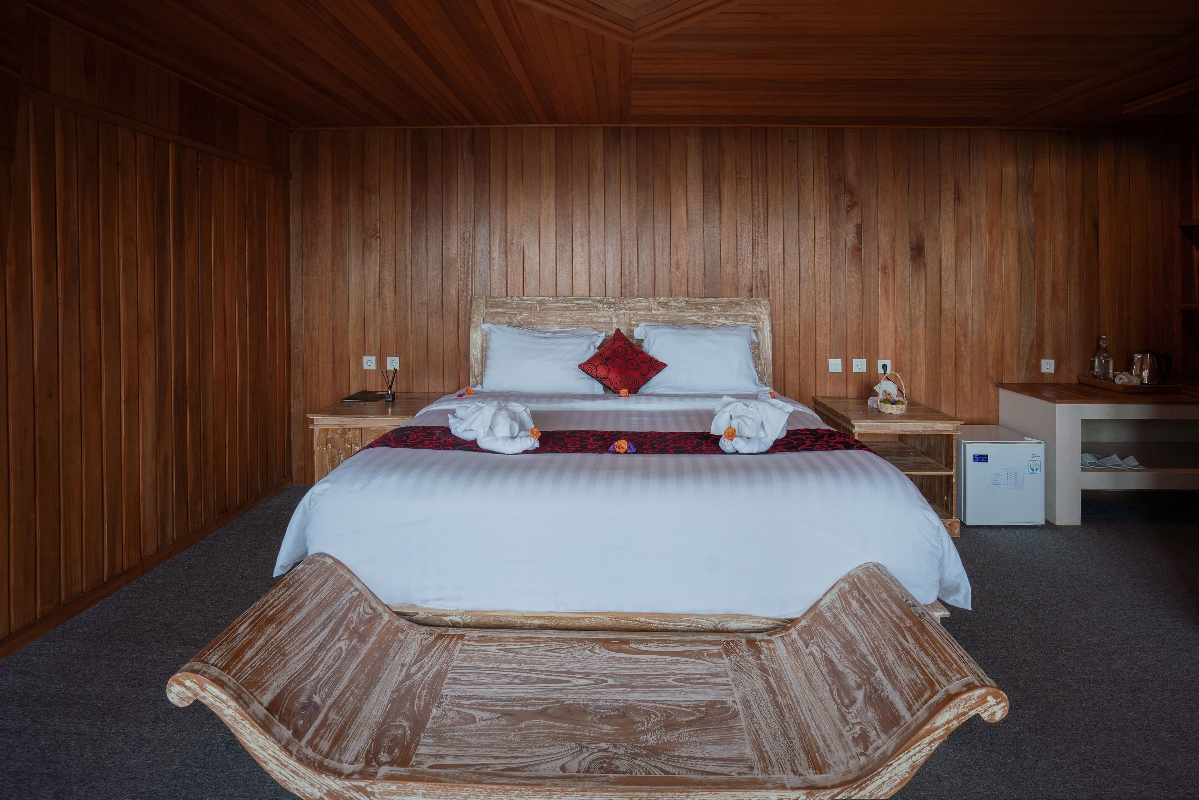 Una habitación con paneles de madera, una cama, dos mesitas de noche, un refrigerador pequeño y un banco.