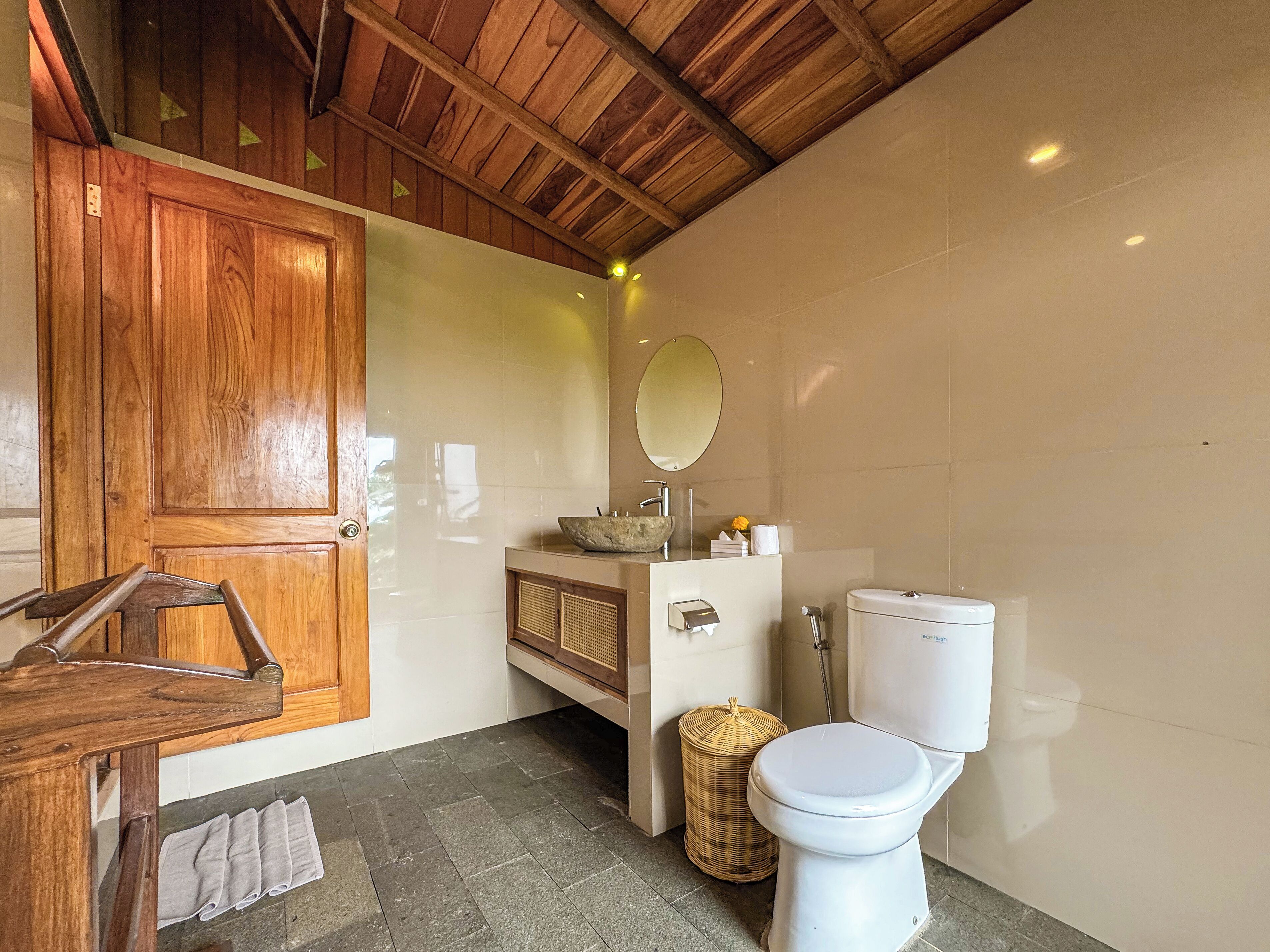 Une salle de bain avec une porte en bois, un lavabo blanc avec un receveur en pierre, un miroir rond, des toilettes et un panier en osier.