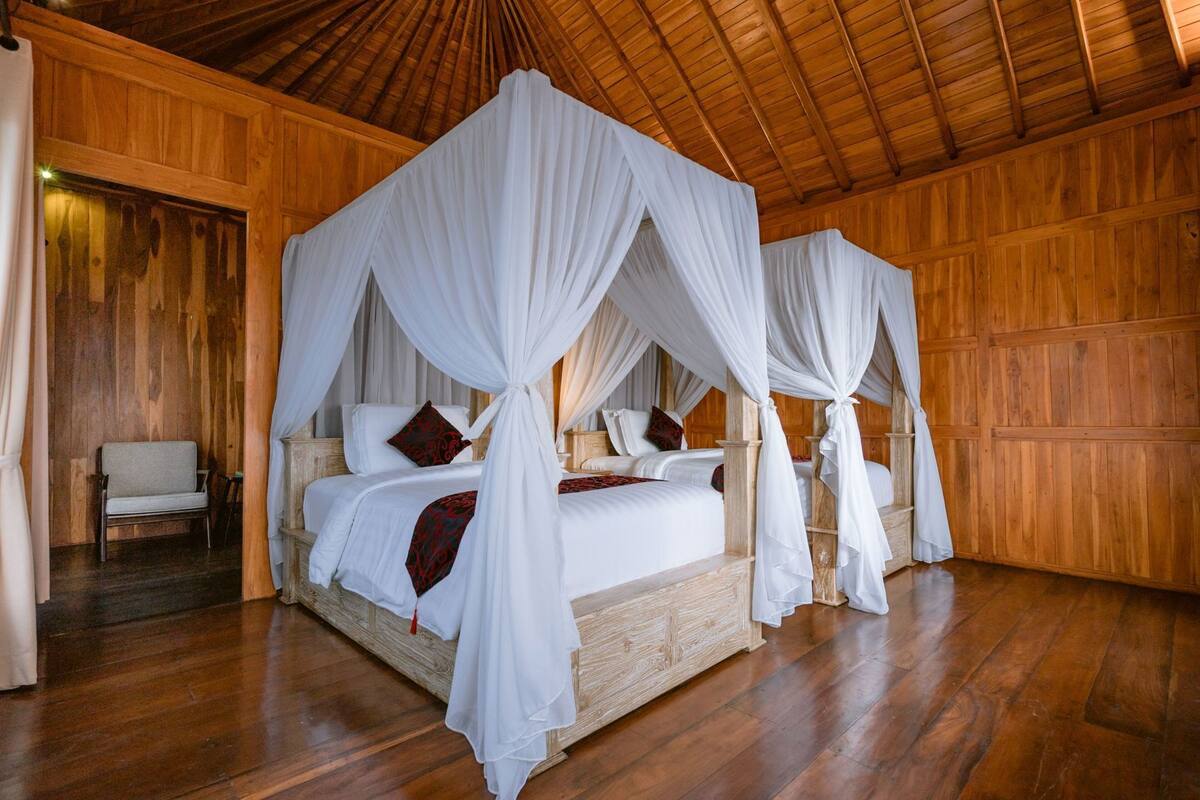 A four-poster canopy bed with white linens and red accents, wooden flooring, and a wooden ceiling.