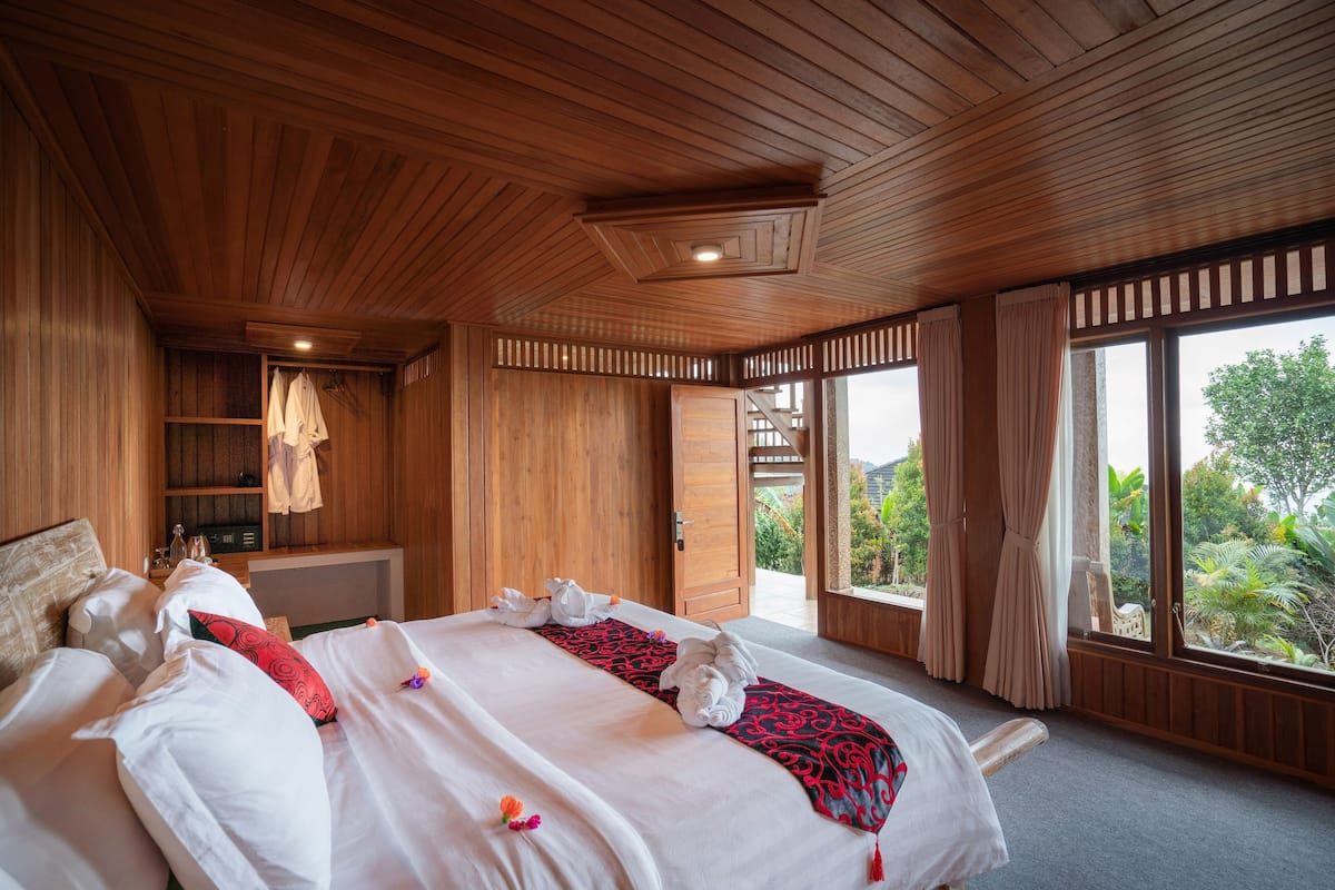 A wooden-paneled bedroom with a large bed, a walk-in closet, and a view of greenery through the windows.
