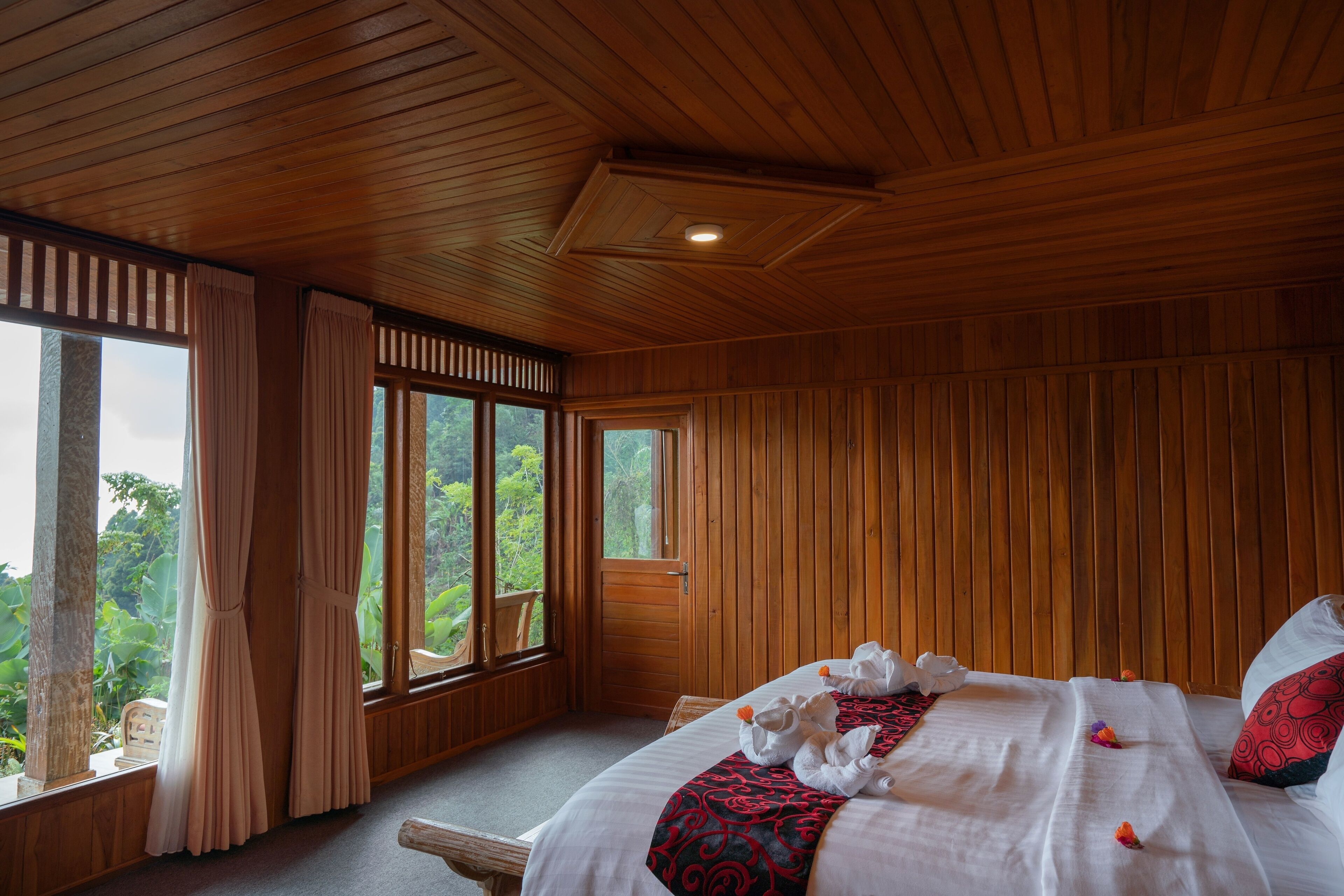 Una habitación con paneles de madera, una cama, ventanas amplias y vistas al verdor exterior.