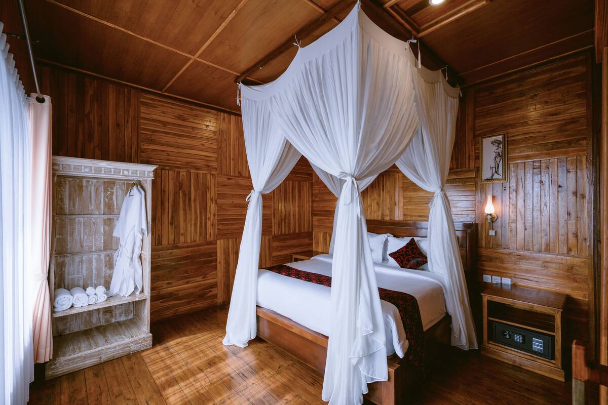 A wooden room with a four-poster bed, a shelf with towels, and a bedside table with a lamp.
