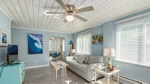 A living room with a ceiling fan, a sofa, a coffee table, and artwork on the walls. - Beautiful downtown Melbourne Beach renovated apartment one block from the beach! (Melbourne Beach)