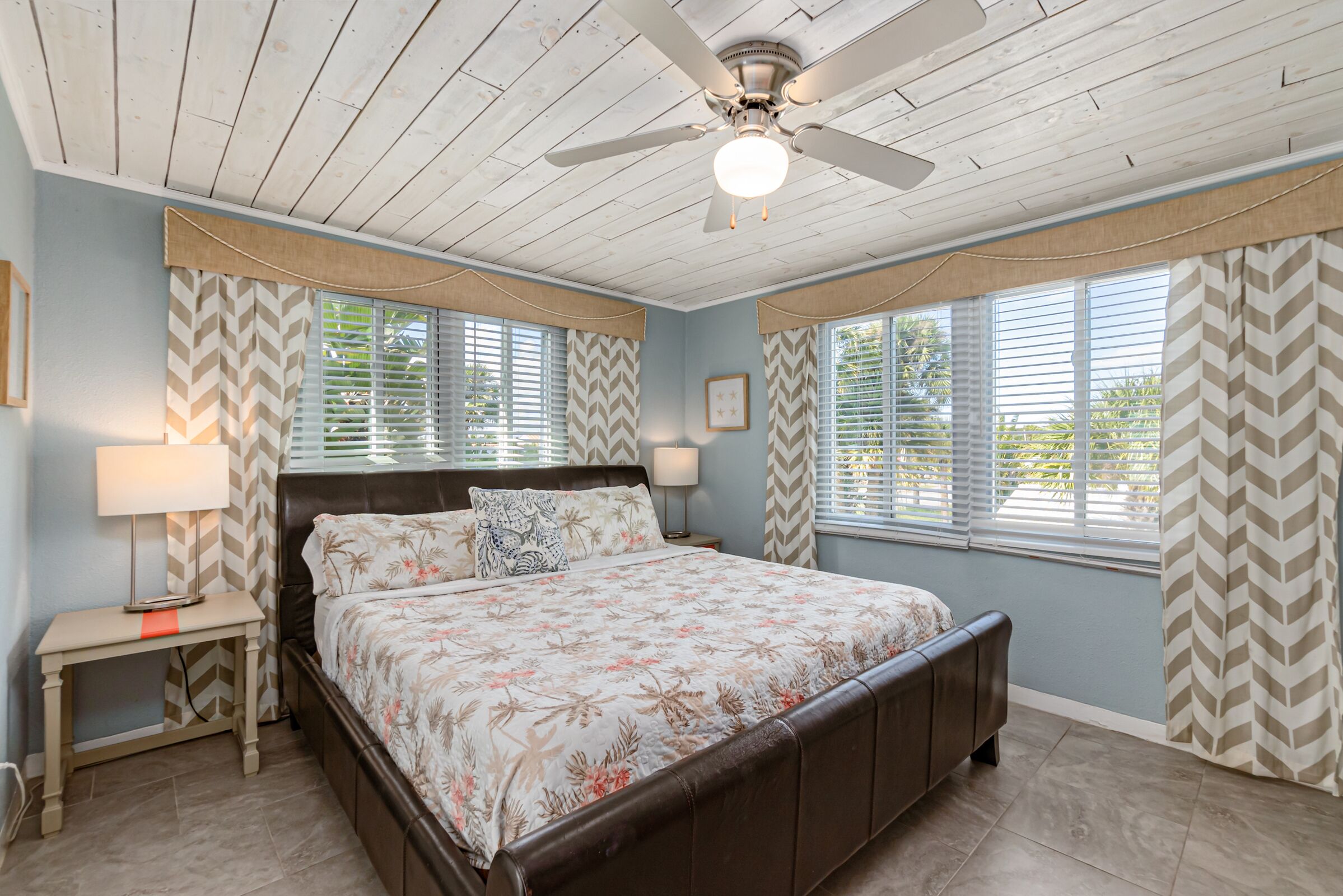 A bedroom with a bed, a ceiling fan, a nightstand, and windows with curtains.