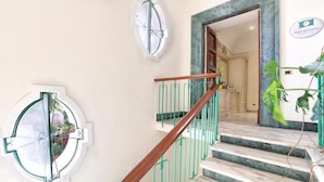 Una escalera con barandilla de madera, pasamanos de metal verde y vista a un cuarto con piso de mármol y una planta.