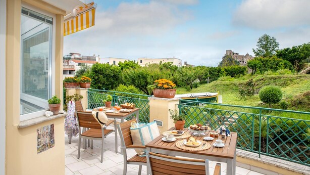 Un balcón con mesa dispuesta para el desayuno, plantas en macetas y vistas a una colina con un castillo.