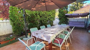 A covered patio with a table and chairs, surrounded by greenery and a white fence.