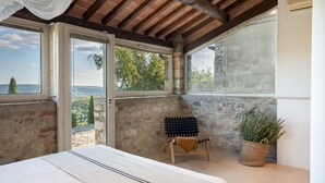 En balkon med stenvægge, en træbænk, en potteplante og en seng med hvidt sengetæppe.