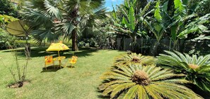 A lush garden with palm trees, a yellow umbrella, and a small table with chairs.