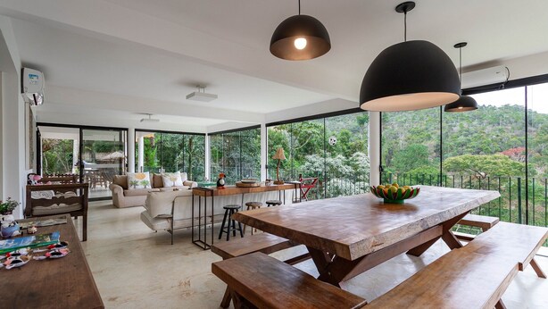 Un comedor espacioso con una gran mesa de madera y bancos, rodeado de amplios ventanales que ofrecen vistas de vegetación exuberante.