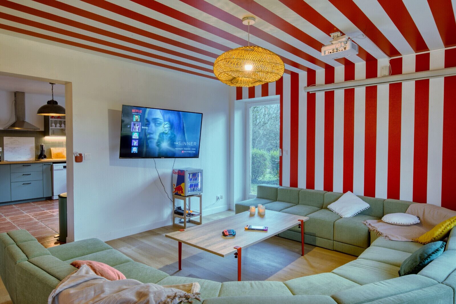 En moderne stue med et stribet rødt og hvidt loft, et fladskærms-tv, en grøn sofa, et træsofabord og et køkkenområde i baggrunden.