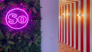 Un mur orné d’un néon représentant le mot « So », entouré de verdure, et un couloir aux murs rayés de rouge et de blanc, agrémenté de luminaires fixés au mur.