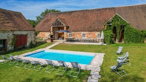 Une piscine avec des chaises longues, un bâtiment en briques doté d’un toit en pente et un espace extérieur couvert aménagé en salon.