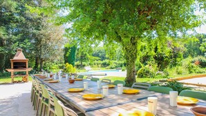 A long wooden dining table set for a meal with chairs, surrounded by lush greenery and a barbecue grill. - So Villa Chasseuserie (89) - Heated pool - Foot - 2h Paris - 45p. (Auxerre)
