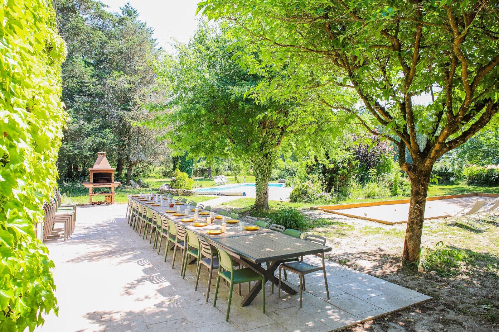 Ein Essbereich im Freien mit einem langen Tisch, der für eine Mahlzeit gedeckt ist, umgeben von Bäumen und einem Pool im Hintergrund.