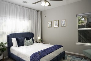 Ein Schlafzimmer mit einem Bett, einem Deckenventilator, einem Fenster mit Vorhängen und gerahmten Bildern an der Wand.