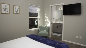Ein Schlafzimmer mit Bett, Fenster, Sessel, Fernseher und einem durchgehenden Türspalt sichtbaren Badezimmer.