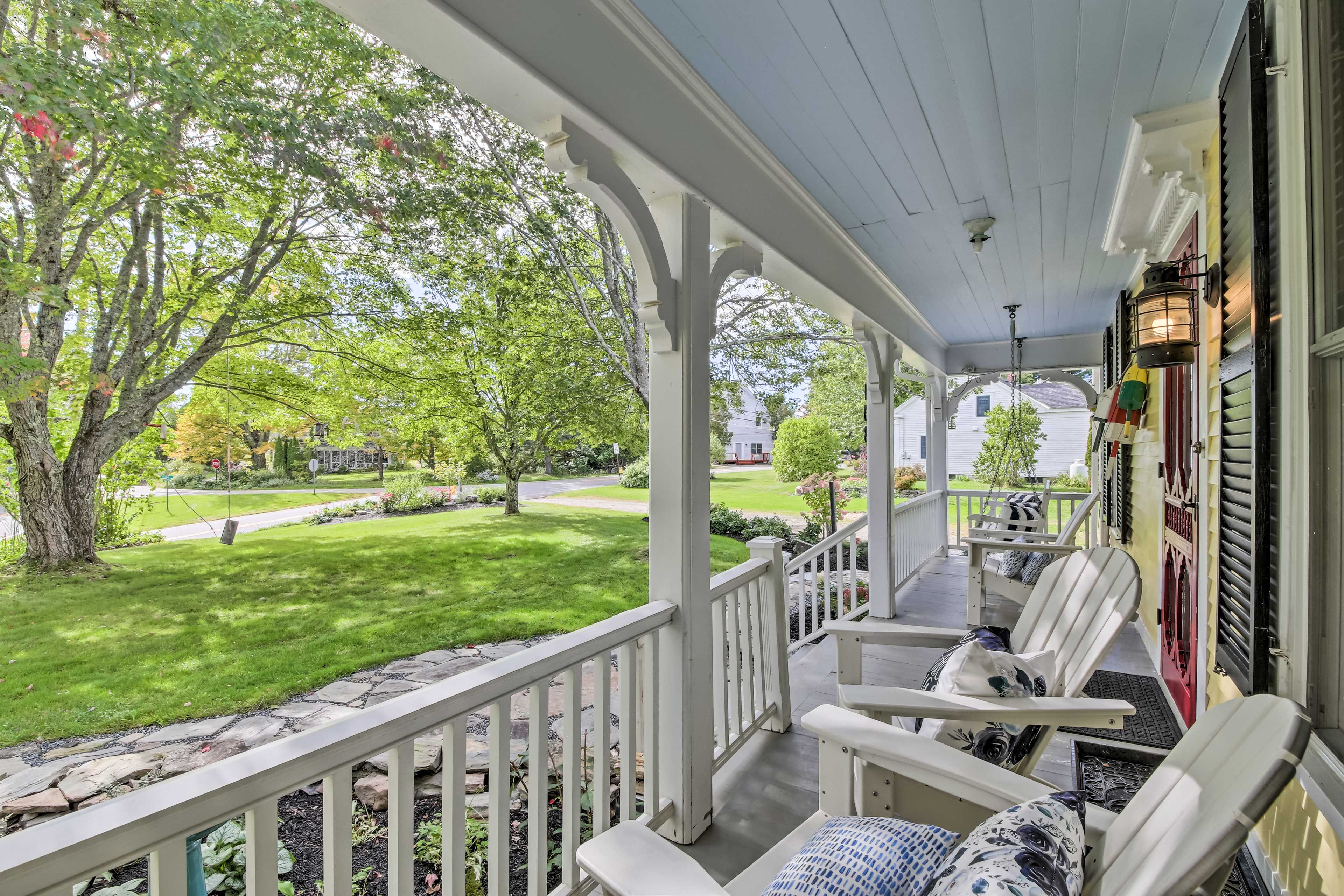 A covered porch with white railings, two white chairs with patterned cushions, and a view of a green lawn and trees.