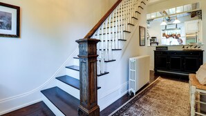 A staircase with a wooden handrail and white balusters, leading to a kitchen area with a white radiator and a hanging light fixture.