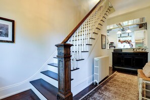 A staircase with a wooden handrail and white balusters, leading to a kitchen area with a white radiator and a hanging light fixture.