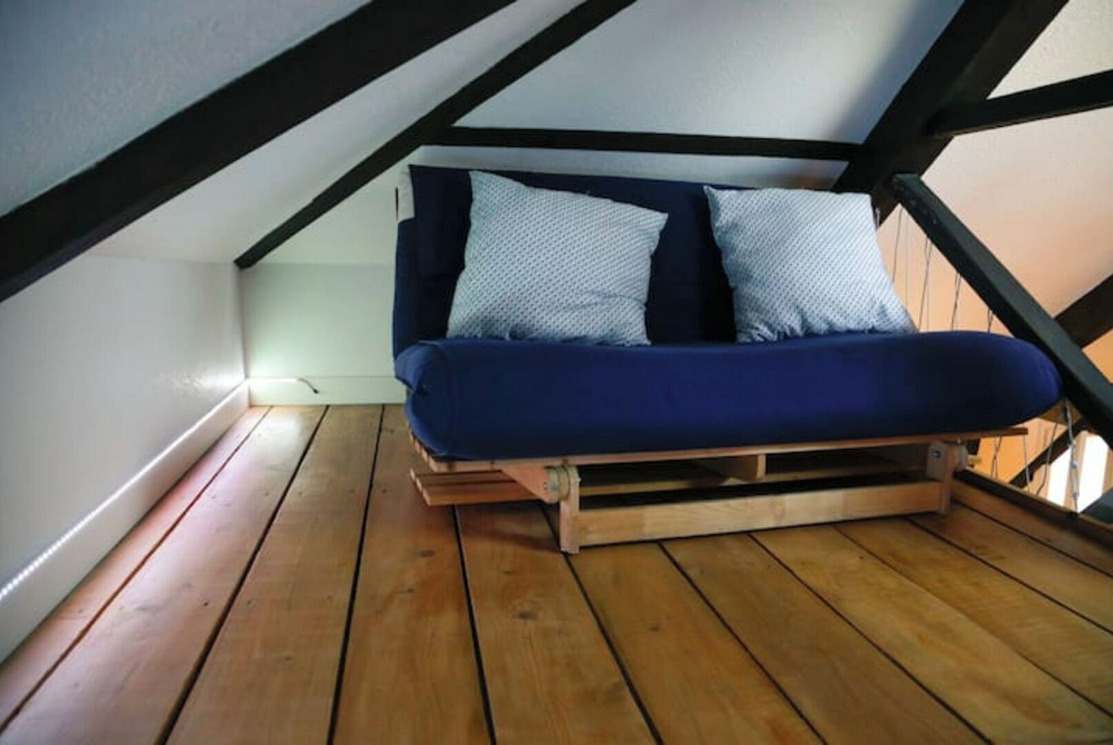 A loft-style bedroom with a wooden floor, a blue bed with patterned pillows, and a sloped ceiling.